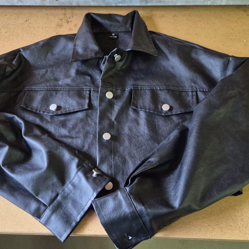 A black leather jacket with a collar, multiple pockets, and silver buttons is laid flat on a brown surface.