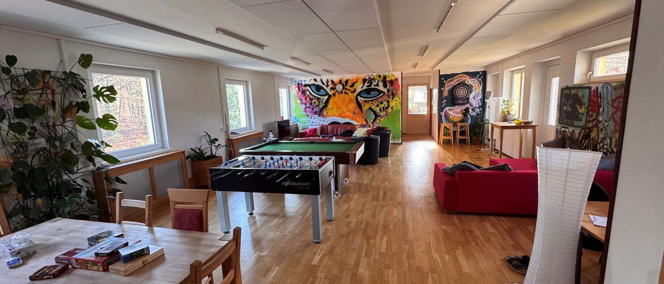 Ein leerer Raum mit Holzboden, ausgestattet mit einem roten Sofa, einem Tischfußballtisch und einem Tisch mit Büchern. Eine farbenfrohe Tigermalerei ziert eine Wand.