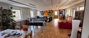 Ein leerer Raum mit Holzboden, ausgestattet mit einem roten Sofa, einem Tischfußballtisch und einem Tisch mit Büchern. Eine farbenfrohe Tigermalerei ziert eine Wand.