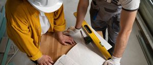 Zwei Männer in Arbeitskleidung, einer in einer gelben Jacke und einem Schutzhelm, der andere in einer Latzhose, untersuchen eine Bauzeichnung auf einem Holztisch. Sie verwenden Werkzeuge und Messinstrumente.