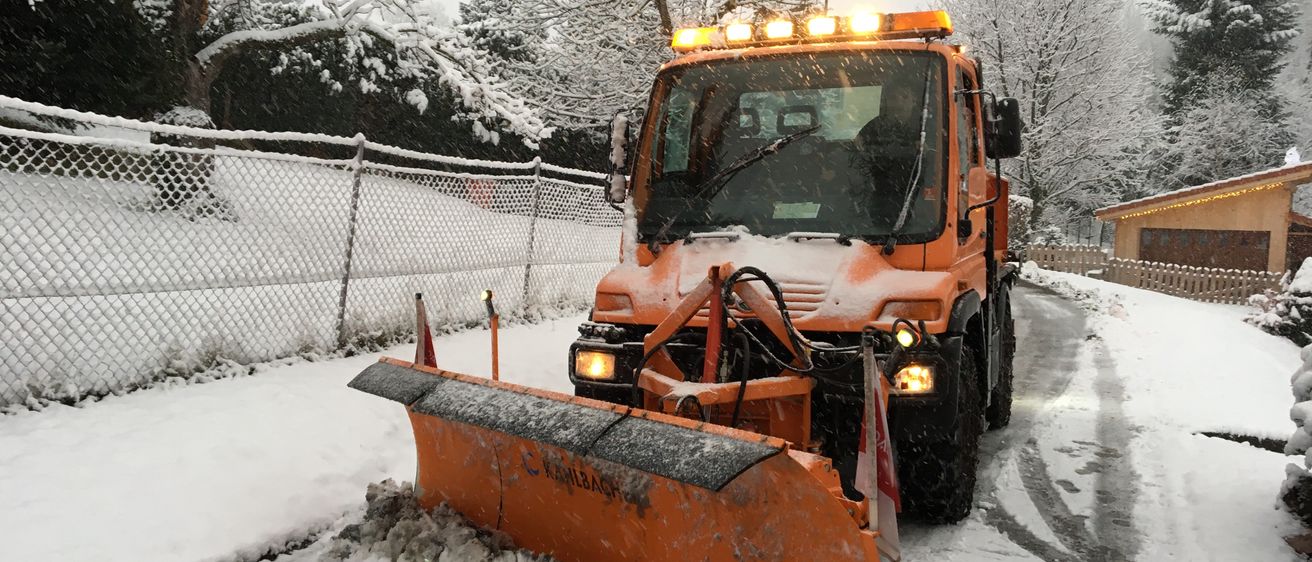 Ein orangefarbener Schneepflug räumt Schnee auf einer schneebedeckten Straße. Dahinter befindet sich ein Maschendrahtzaun, und im Hintergrund sind Bäume mit Schnee darauf.