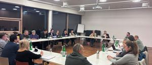 A meeting is taking place in a conference room. People are seated around tables, and some are standing. There are bottles, laptops, and papers on the tables. The room has white walls, a projector, and large windows.