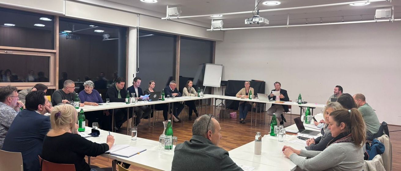 Eine Besprechung findet in einem Konferenzraum statt. Die Menschen sitzen um Tische herum, einige stehen. Auf den Tischen befinden sich Flaschen, Laptops und Papiere. Der Raum hat weiße Wände, einen Projektor und große Fenster.