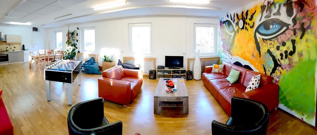 Ein geräumiges Wohnzimmer mit einem roten Sofa, einem Couchtisch und einer großen Wandkunst. Es gibt einen Tischfußball, einen Fernseher und Lautsprecher.