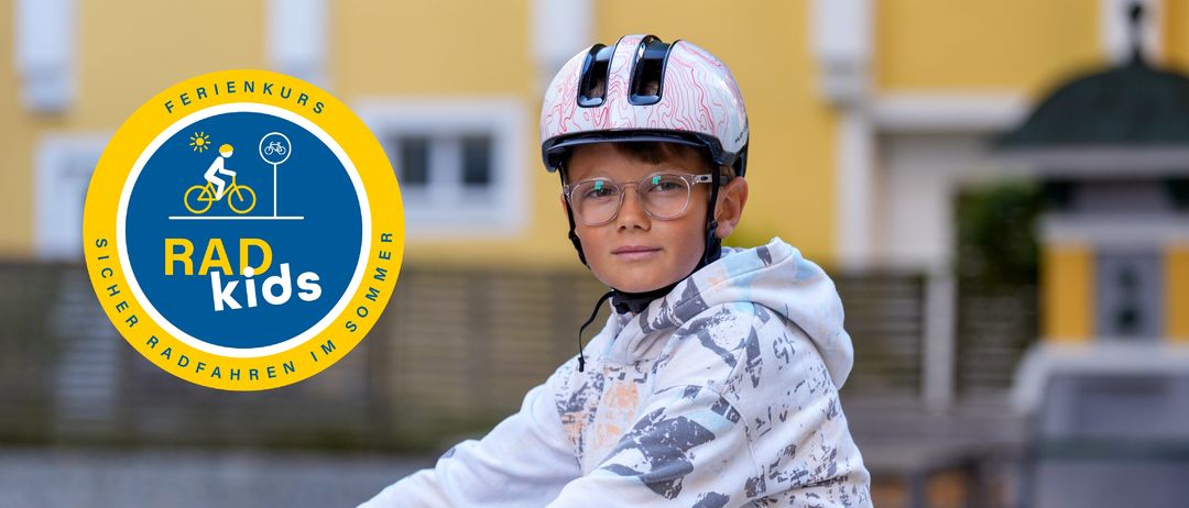 Ein Junge fährt ein Fahrrad und trägt einen Helm, eine Brille und eine Kapuzenjacke mit einem farbenfrohen Design. Hinter ihm steht ein gelbes Gebäude mit einem Logo.