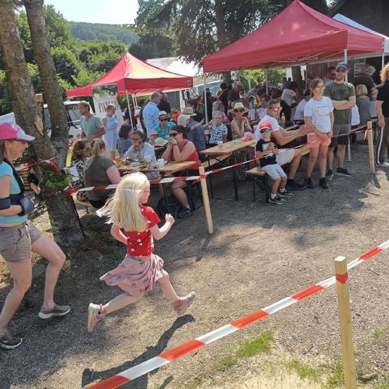 Eine Menschenmenge versammelt sich bei einer Outdoor-Veranstaltung unter roten Zelten. Einige sitzen an Tischen, andere stehen, und ein Mädchen rennt an einer Barriere vorbei. Bäume und Grün umgeben das Gelände.
