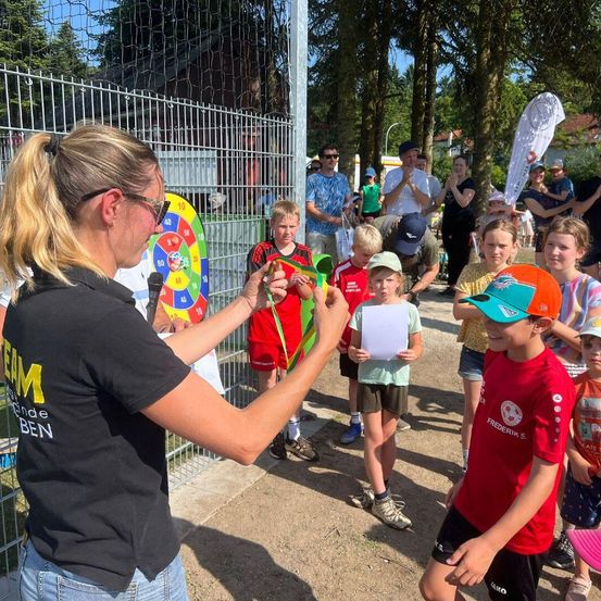 Eine Frau in einem schwarzen Shirt überreicht einem Jungen in einem roten Shirt eine Medaille bei einer Veranstaltung im Freien. Dahinter beobachten eine Gruppe von Kindern und Erwachsenen. Ein Metallzaun und ein Dartboard sind im Hintergrund.