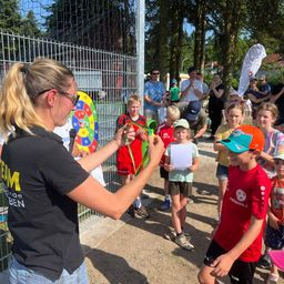 Eine Frau in einem schwarzen Shirt überreicht einem Jungen in einem roten Shirt eine Medaille bei einer Veranstaltung im Freien. Dahinter beobachten eine Gruppe von Kindern und Erwachsenen. Ein Metallzaun und ein Dartboard sind im Hintergrund.