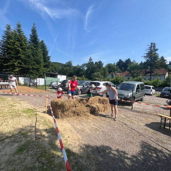 Eine Gruppe von Menschen, einschließlich Kindern, spielt auf einem Feld mit Heuballen. Eine Frau hilft einem Kind, einen Heuballen zu erklimmen. Einige Autos sind auf einem Parkplatz geparkt.
