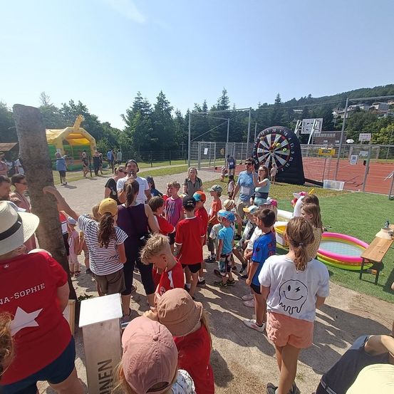 Eine Gruppe von Kindern und Erwachsenen versammelt sich um ein Dartboard, ein Hüpfhaus und einen kleinen Pool. Die Veranstaltung findet im Freien an einem sonnigen Tag statt, mit einigen Bäumen und einem Gebäude in der Ferne.
