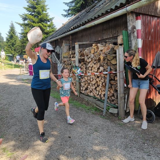 Eine Frau mit der Nummer 15 läuft mit einem Mädchen. Eine Frau mit einem Buch steht neben einem Holzstapel.