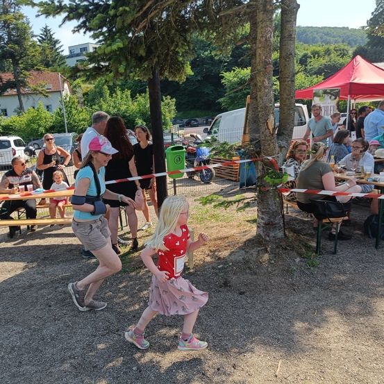 Eine Gruppe von Menschen, darunter eine Frau mit einer Armstütze, geht in der Nähe eines Baumes, während im Hintergrund ein rotes Zelt steht. Einige Leute sitzen an Tischen mit Essen.