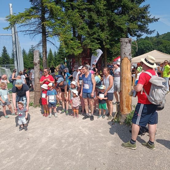Eine Gruppe von Menschen, einschließlich Kindern, steht in einer Schlange unter Bäumen. Einige tragen Hüte, Brillen und Turnschuhe. Bäume und Zelte sind im Hintergrund.