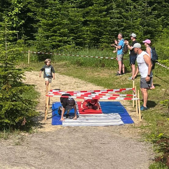 Kinder machen ein Hindernisparcours auf einem Lehmweg, umgeben von hohen Bäumen. Ein Junge läuft auf eine rote Matte zu, während Erwachsene zusehen.