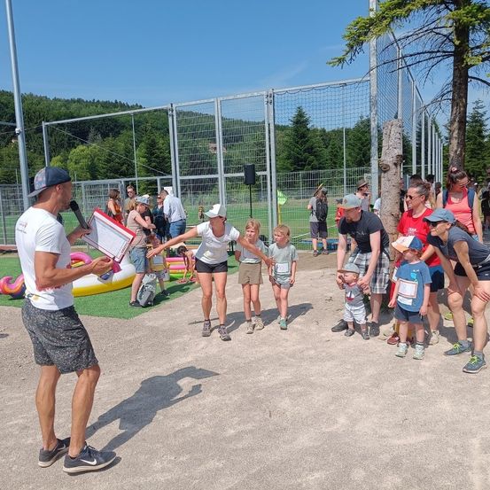 Eine Gruppe von Menschen versammelt sich in einem umzäunten Außenbereich. Ein Mann hält ein Mikrofon und Papier und führt eine Reihe von Kindern und Erwachsenen an. Dahinter sind aufblasbare Gegenstände und ein Metallzaun zu sehen, mit Bäumen und blauem Himmel im Hintergrund.