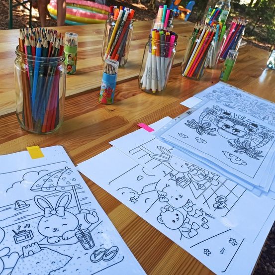 Ein Holztisch hat Gläser mit Buntstiften und Markern, Malblätter und andere Gegenstände. Die Malblätter haben Zeichnungen von einem Hasen, einem Bären und einer Blume.