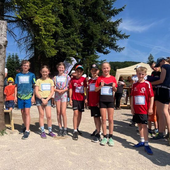Eine Gruppe von Kindern steht draußen unter Bäumen. Sie tragen alle Sportshirts und Shorts. Sie halten ein Zertifikat in der Hand und tragen Turnschuhe. Sie scheinen Gewinner eines Wettbewerbs zu sein.