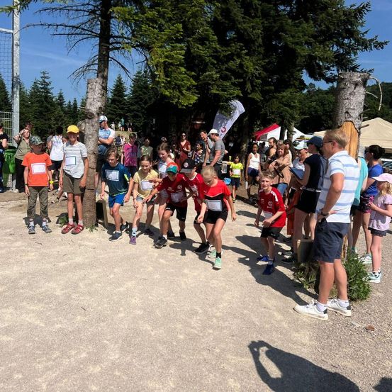 Eine Gruppe junger Athleten mit Startnummern steht auf einer Laufbahn in einem Park, Zuschauer beobachten.