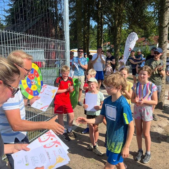 Eine Gruppe von Kindern und Erwachsenen versammelt sich draußen. Ein Mann hält Urkunden, während andere Papiere halten. Sie stehen vor einem Zaun, mit einer Fahne auf der rechten Seite.