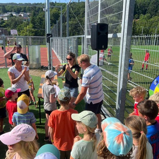 Eine Gruppe von Kindern und Erwachsenen versammelt sich auf einem Feld mit Zaun. Ein erwachsener Mann und eine Frau stehen neben einem Lautsprecher und sprechen zu den Kindern. Im Hintergrund spielen Menschen Fußball.