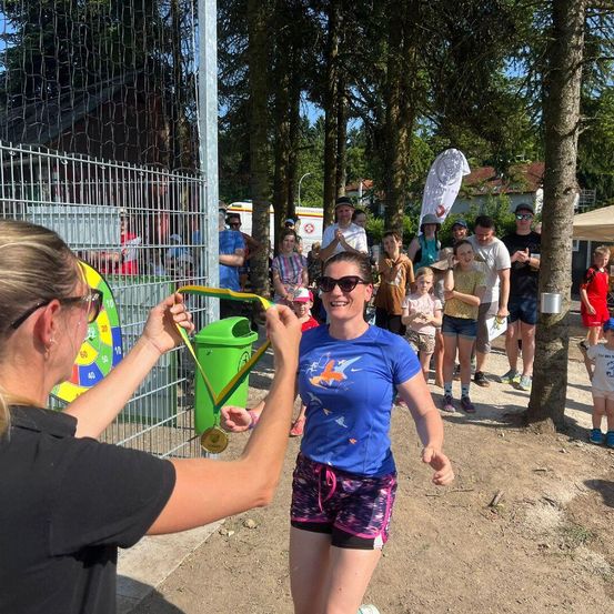 Eine Frau wird für einen Marathon mit einer Medaille ausgezeichnet, neben einem Mülleimer stehend, während mehrere Menschen im Hintergrund zusehen.