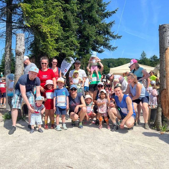 Eine Gruppe von Menschen, einschließlich Erwachsener und Kindern, posiert für ein Foto im Freien. Einige tragen Hüte, Brillen und Turnschuhe, einer trägt ein Kind. Bäume und ein Zelt sind im Hintergrund.