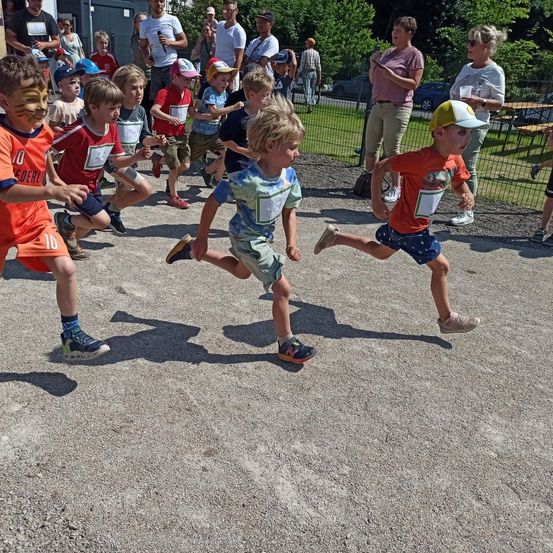 Kinder nehmen an einem Rennen auf einem Kiesweg teil, tragen nummerierte Trikots und Mützen. Zuschauer stehen hinter einem Zaun und beobachten die Veranstaltung.