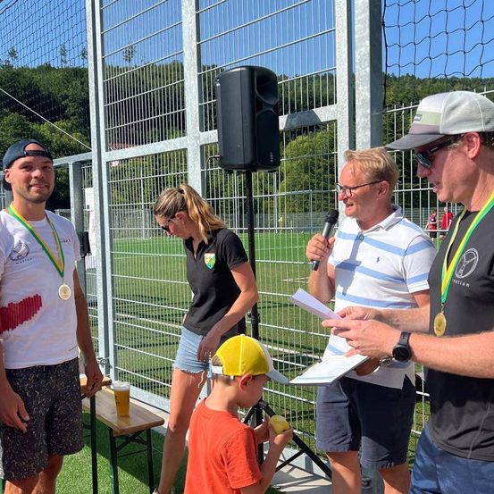 Ein Mann mit einer Medaille steht mit einem Bierglas in der Nähe eines Zauns. Eine Frau steht hinter ihm. Ein Mann mit einem Mikrofon spricht mit einem Mann, der eine Mütze trägt. Ein Junge schaut einen Apfel an.