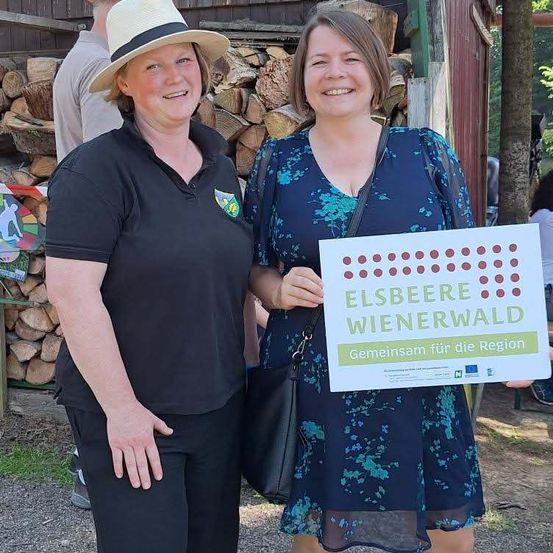 Zwei Frauen stehen lächelnd vor einem Hintergrund aus gestapeltem Holz, eine hält ein Schild zur Förderung von Elsbeere Wienerwald.
