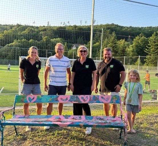 Fünf Erwachsene und ein Kind stehen vor einer dekorierten Bank auf einem Fußballfeld mit einem Tornetz.