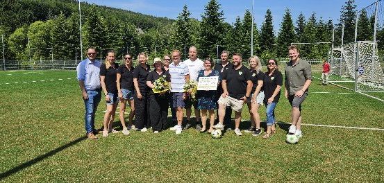 Eine Gruppe von Menschen steht auf einem Fußballfeld mit Fußballbällen und hält eine Urkunde. Sie tragen verschiedene Kleidungsstücke und Sonnenbrillen. Das Feld ist von Bäumen und einem Zaun umgeben.