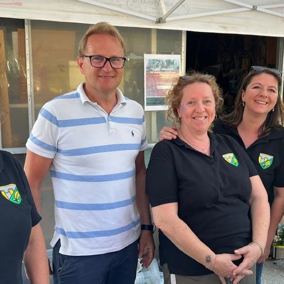 Vier Personen stehen unter einem weißen Zelt. Ein Mann trägt ein gestreiftes Hemd, während drei Frauen schwarze Shirts tragen. Sie alle lächeln in die Kamera. Hinter ihnen spiegelt ein Fenster das Zelt.