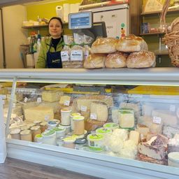 Eine Frau steht hinter einer Käsetheke in einem Laden und zeigt verschiedene Käsesorten und Brot. Im Hintergrund befinden sich Regale und ein Korb.