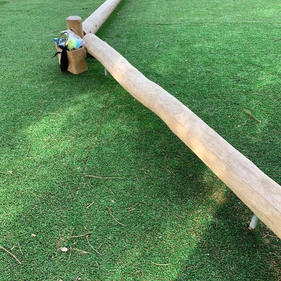 Ein großer Holzbalken liegt auf grünem Kunstrasen, an dem ein kleiner Korb befestigt ist, der verschiedene Gegenstände enthält.