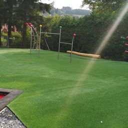 Ein Kinderspielplatz mit Kunstrasen und Spielgeräten. An Stangen sind verschiedenfarbige Bälle befestigt. In der Mitte befindet sich eine Holzbrücke.