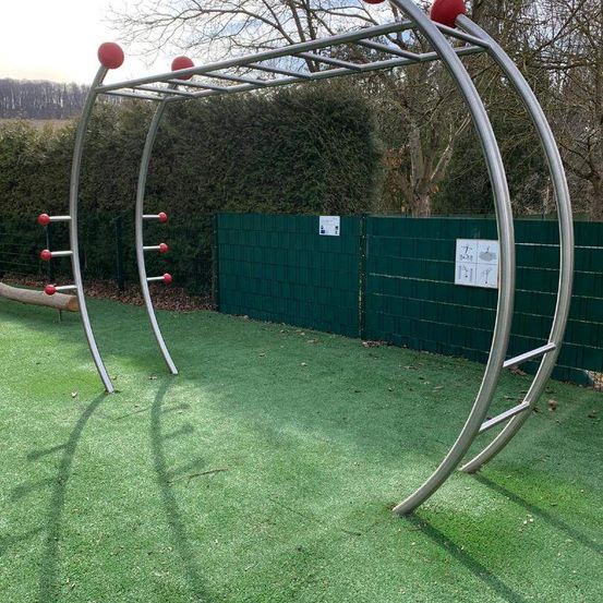 Ein Metall-Klettergerüst mit roten Kugeln steht auf einem grünen Rasen. Dahinter befindet sich ein grüner Zaun.