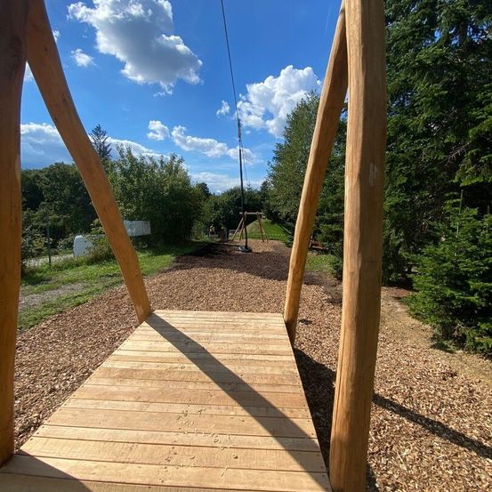Eine Holzbrücke mit Blick auf einen bewölkten Himmel und ein Spielplatz in der Ferne. Bäume und Pflanzen umgeben das Gelände.