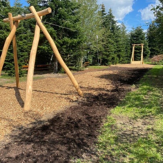 Ein Außenspielplatz mit Holzschaukeln und einem mit Mulch bedeckten Weg. Hohe Bäume umgeben den Bereich, und eine Bank ist im Hintergrund zu sehen.