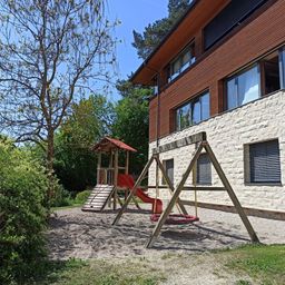 Ein modernes Gebäude mit Holzverkleidung und Glasfenstern verfügt über einen Kinderspielplatz mit Rutsche und Schaukel. Bäume und Büsche umgeben das Gelände.