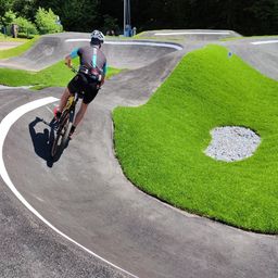 Ein Radfahrer mit Helm fährt eine abschüssige Asphaltbahn hinunter, mit saftigem grünem Gras auf beiden Seiten. Die Strecke hat Kurven und weiße Markierungen. In der Ferne befinden sich weitere Strecken und Grünflächen.