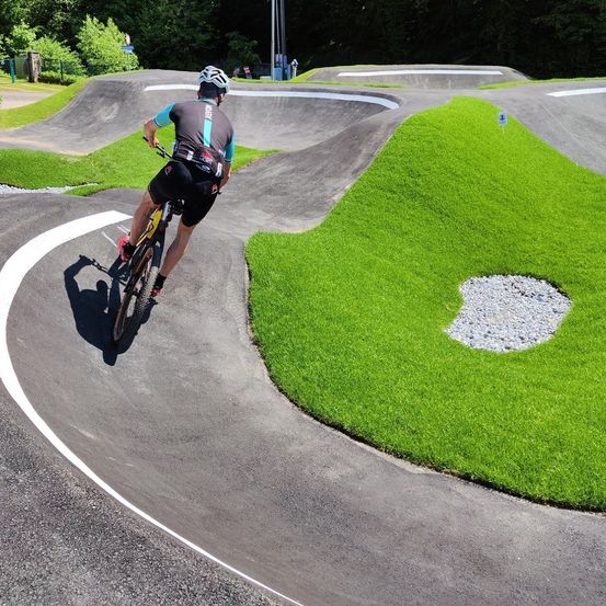 Ein Radfahrer mit Helm fährt eine abschüssige Asphaltbahn hinunter, mit saftigem grünem Gras auf beiden Seiten. Die Strecke hat Kurven und weiße Markierungen. In der Ferne befinden sich weitere Strecken und Grünflächen.