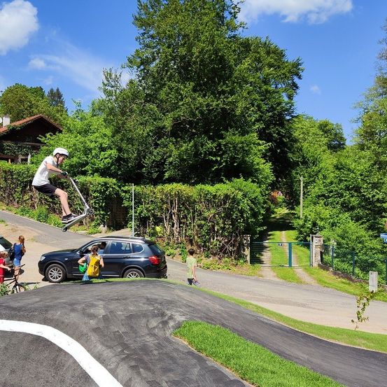 Eine Person macht einen Trick mit einem Roller, während Leute in der Nähe beobachten, ein geparktes Auto auf einer grün umrahmten abschüssigen Straße.
