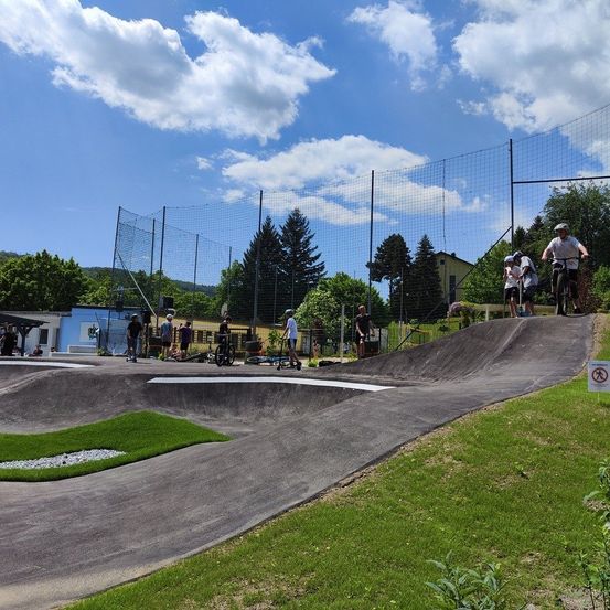 Ein Skatepark mit einigen Leuten, die an einem sonnigen Tag Fahrrad fahren und Skateboard fahren. Es gibt zwei Rampen und einen Rasenbereich an der Seite.