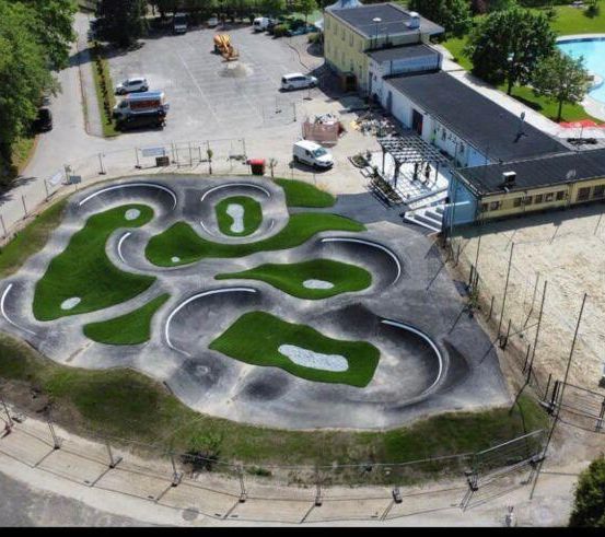 Ein Luftbild eines Skateparks mit grünen Rampen, umgeben von einem Zaun. In der Nähe sind ein Pool und mehrere Gebäude zu sehen. Autos sind in einem Parkplatz neben dem Skatepark geparkt.