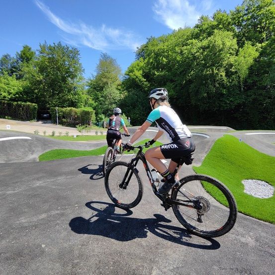 Zwei Radfahrer fahren auf einem Radweg, umgeben von viel Grün, mit einer malerischen Aussicht auf Bäume und blauen Himmel.