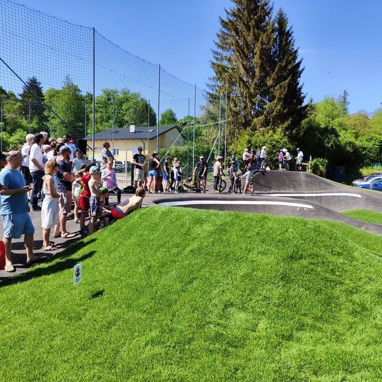 Eine Gruppe von Menschen beobachtet einen BMX-Wettkampf an einem sonnigen Tag, einige auf der Strecke und andere stehend.