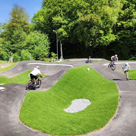 Mehrere Radfahrer fahren auf einer Erdstrecke mit Kurven und Rampen, umgeben von Grün. Bäume säumen die Ränder der Strecke.