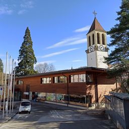 Ein modernes Gebäude mit Holzfassade steht vor einem Turm mit einer Uhr. Der Turm hat einen Spitzhelm und ein Kreuz auf der Spitze. An den Seiten des Turms stehen Bäume. Ein weißes Auto ist vor dem Gebäude geparkt.