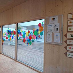 Ein Raum mit Holzwänden und -böden hat ein großes Fenster mit bunten Kreisen darauf. Eine Pinnwand mit Papieren ist an der Wand befestigt.