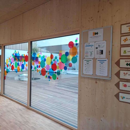 Ein Raum mit Holzwänden und -böden hat ein großes Fenster mit bunten Kreisen darauf. Eine Pinnwand mit Papieren ist an der Wand befestigt.